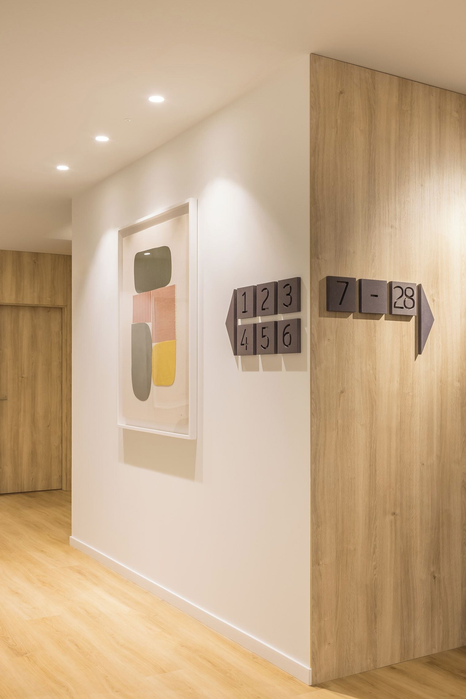 Un pasillo con suelo de madera, una obra de arte abstracta enmarcadas y señalización direccional con números.