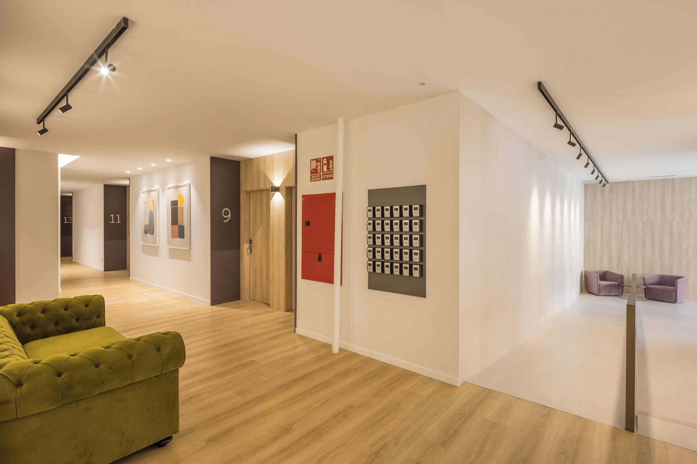 Un vestíbulo de hotel moderno con un sofá verde, suelo de madera, una puerta roja y un sistema de tarjetas de acceso montado en la pared.