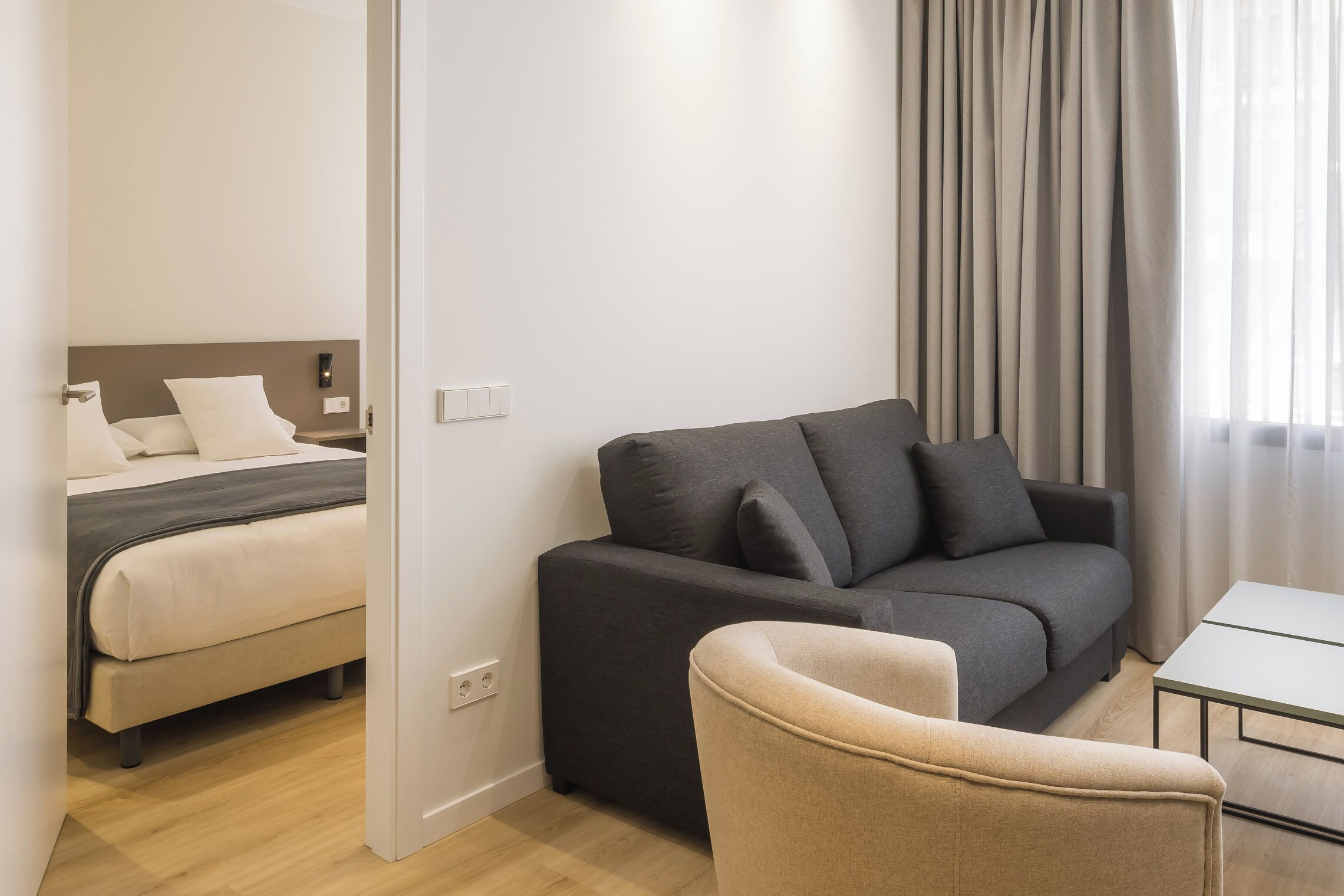 Een moderne hotelkamer met een bed, een donkergrijze bank, een beige fauteuil en een klein wit tafeltje.