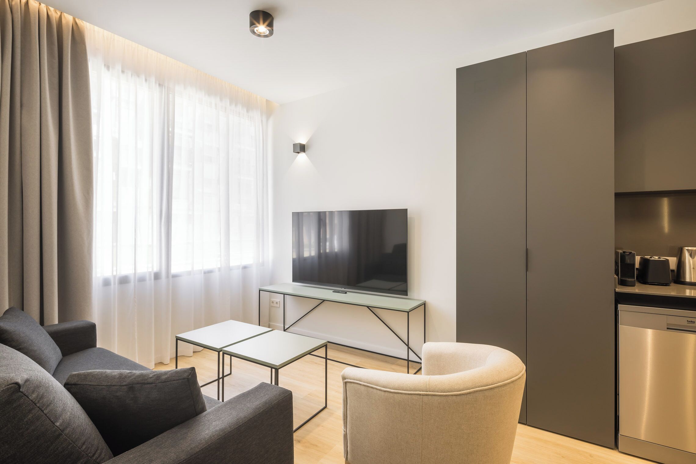 A modern living room with a sofa, a TV on a glass table, and a kitchen area with a dishwasher.
