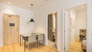 A modern, well-lit room with a wooden floor, a dining table with chairs, a pendant light, and a door leading to a bathroom.