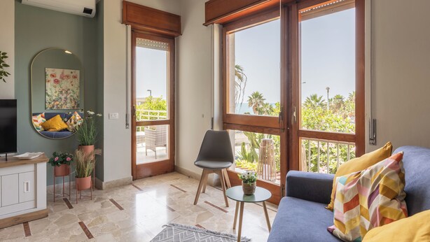 Ein modernes Wohnzimmer mit einer Couch, einem Sessel, einem kleinen Tisch und einem Fernseher. Große Fenster bieten Blick ins Grüne.