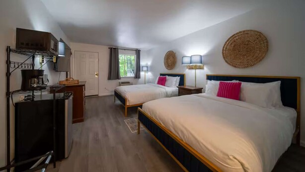 A hotel room with two beds, a kitchenette, a window with curtains, and a round woven wall hanging.
