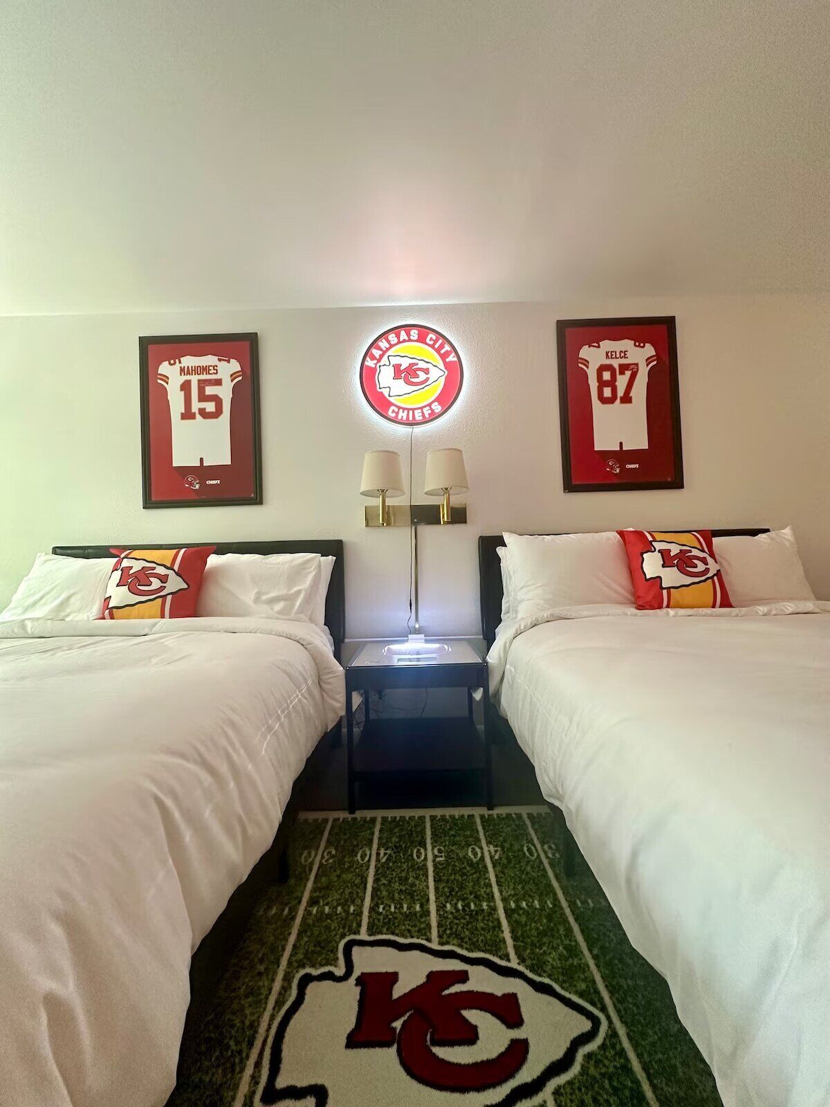 Habitación de hotel con dos camas, cada una con ropa de cama y almohadas con el logo de los Kansas City Chiefs. En la pared, hay camisetas enmarcadas, un logo redondo sobre las camas y una mesita con lámpara entre ellas.
