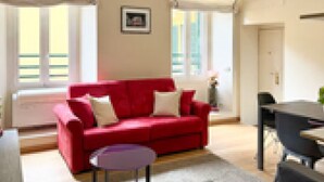 Un sofá rojo en una habitación con una ventana, una mesita redonda y una puerta.