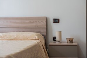 Un lit bien fait, avec une tête de lit en bois, une table de chevet avec une lampe et un petit objet décoratif, et un interrupteur fixé au mur.