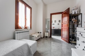 Une chambre à coucher comprenant un lit, une commode, une table de chevet, un radiateur, une fenêtre avec des rideaux et une porte donnant sur une autre pièce.