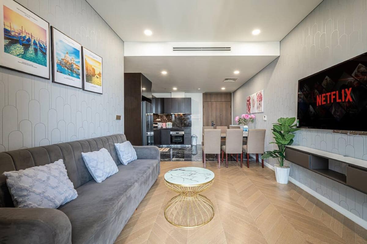 A modern living room with a grey sofa, a round coffee table, a TV on the wall, and a kitchen in the background.