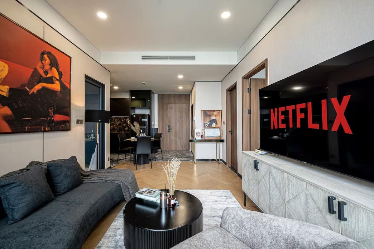 A modern living room with a large flat-screen TV displaying the Netflix logo, a grey sofa, a coffee table, and a dining area in the background.
