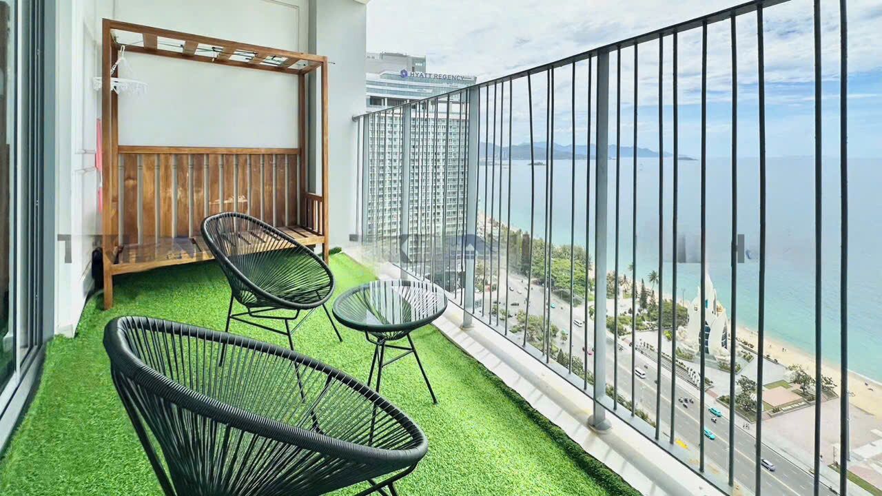 A balcony with artificial grass, two black wicker chairs, and a view of a cityscape and beach.