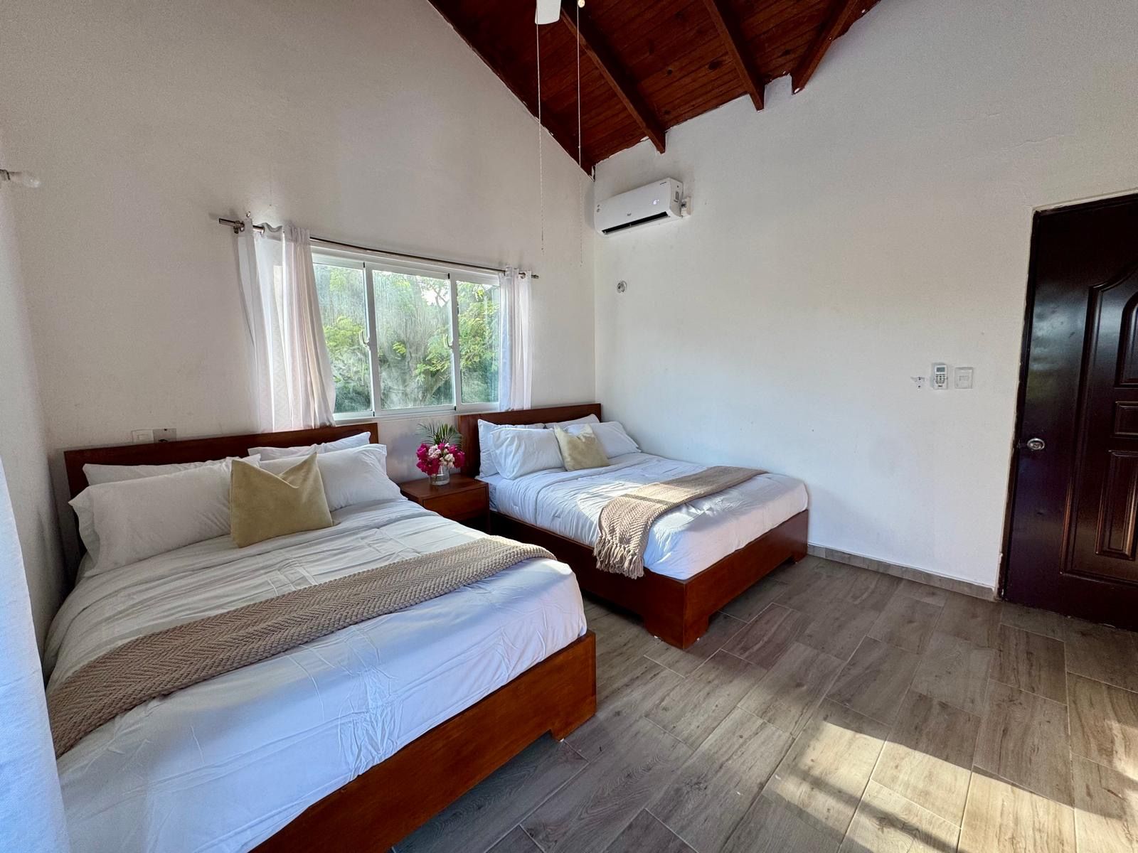Una habitación con dos camas, una ventana con cortinas, un equipo de aire acondicionado y una puerta de madera.