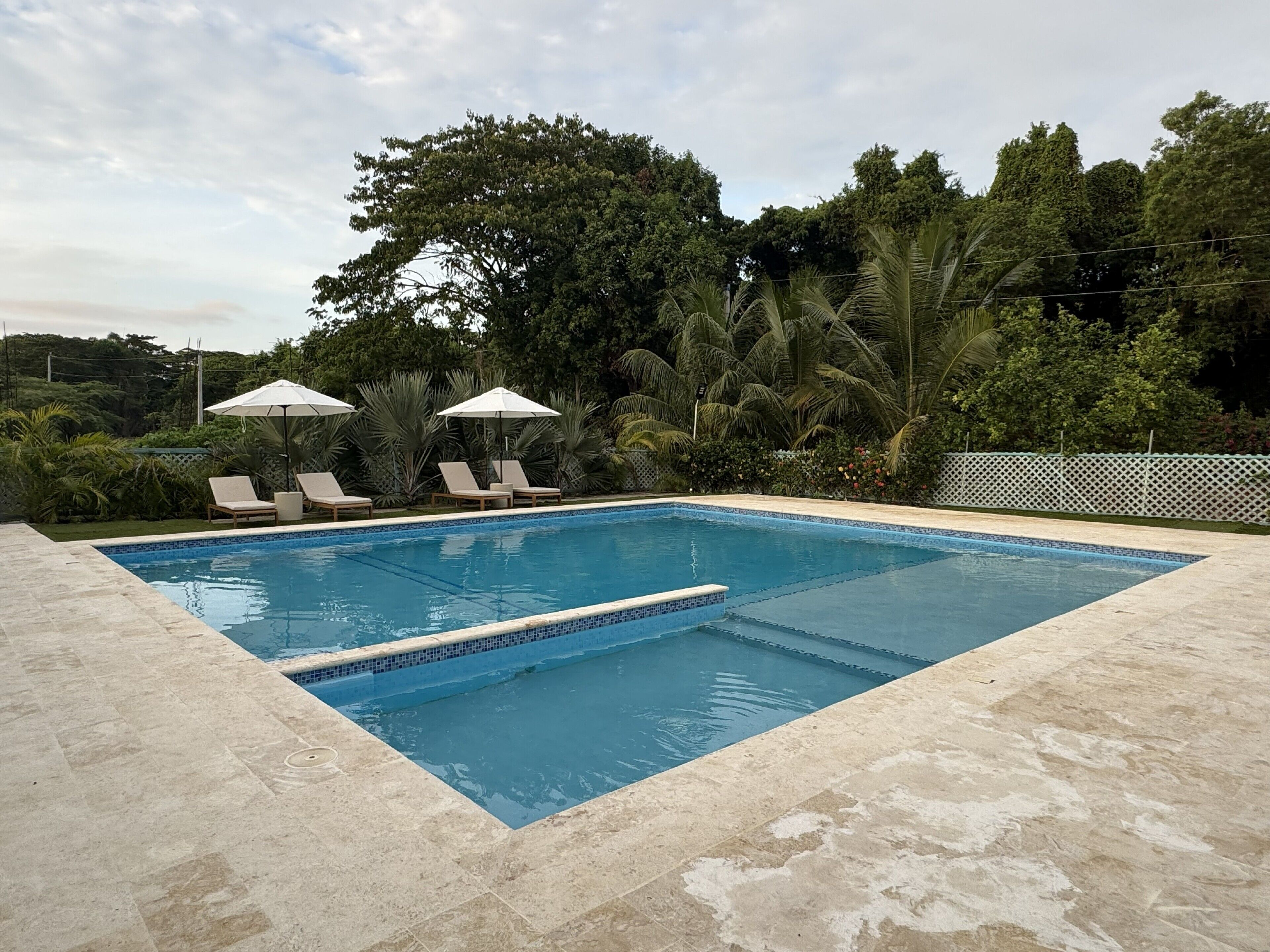 Ein rechteckiger Pool mit gefliestem Rand, umgeben von einer Steindeckfläche. Am Poolufer stehen Liegestühle und Sonnenschirme.