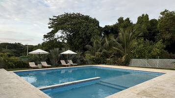 Ein rechteckiger Pool mit gefliestem Rand, umgeben von einer Steindeckfläche. Am Poolufer stehen Liegestühle und Sonnenschirme.