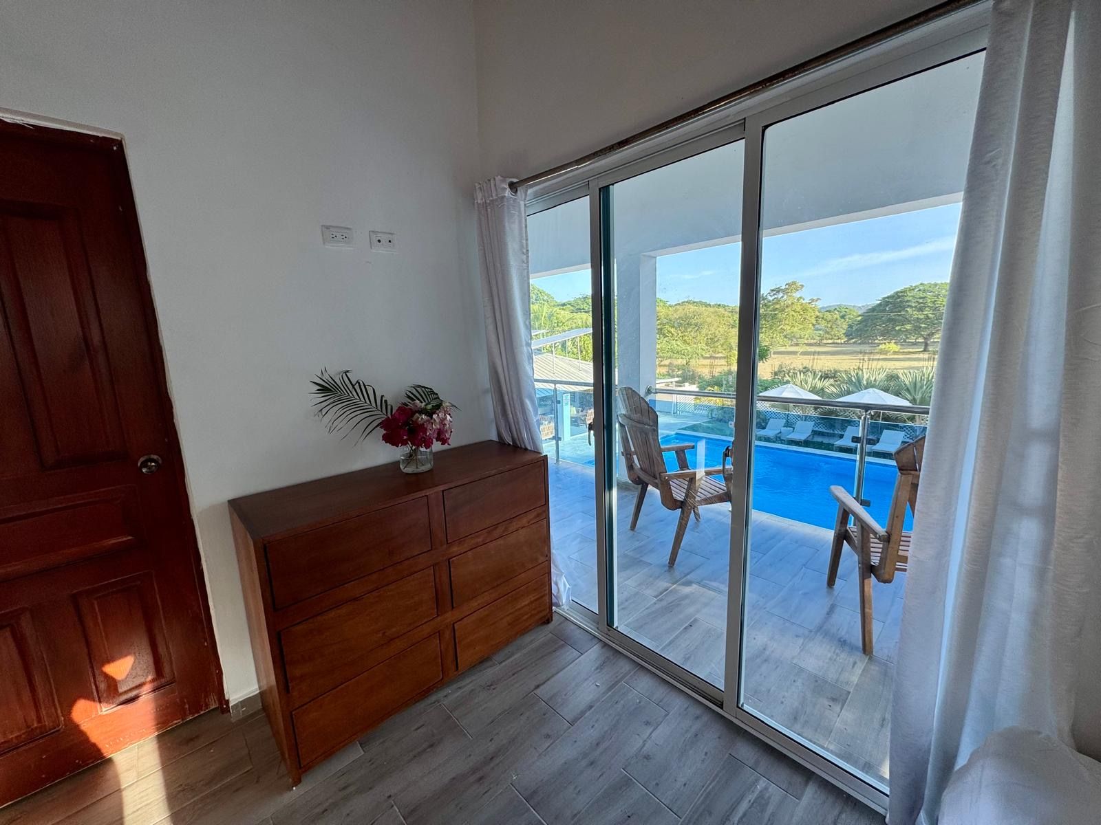 Una habitación con un aparador de madera, puerta de vidrio que da a un balcón con piscina y sillas.