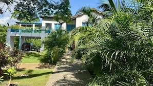 Ein zweistöckiges Haus mit einer Veranda, umgeben von üppiger Vegetation und blauem Himmel.