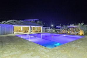 Une piscine avec une terrasse carrelée, un espace de détente couvert avec des chaises et des tables, et un bâtiment doté d’un toit en pente.