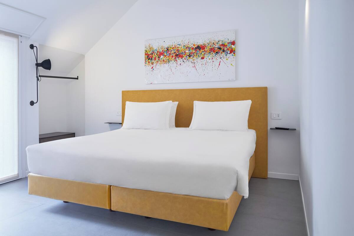 Un dormitorio moderno con cama, ropa de cama blanca, cabecera de madera, una obra de arte colgada en la pared y una mesita de noche.