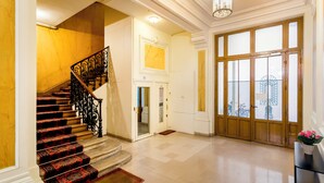 Un magnifique escalier orné de balustrades travaillées, une grande porte en bois avec des panneaux de verre, et une vue sur l’extérieur à travers la porte.
