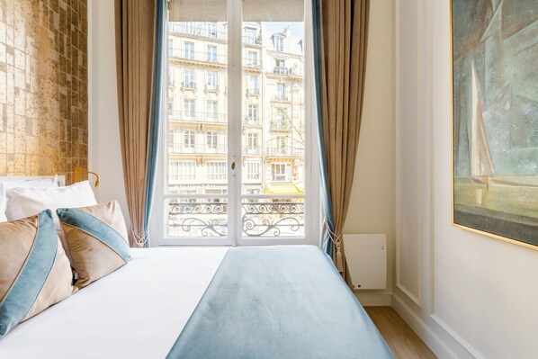 A hotel room with a bed, curtains, a window with a view of buildings, and a painting on the wall. - Merveil Luxury Suite - Louvre Marcel III (Paris)