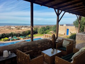 A covered outdoor seating area with a pool, wicker furniture, and a view of the sea.