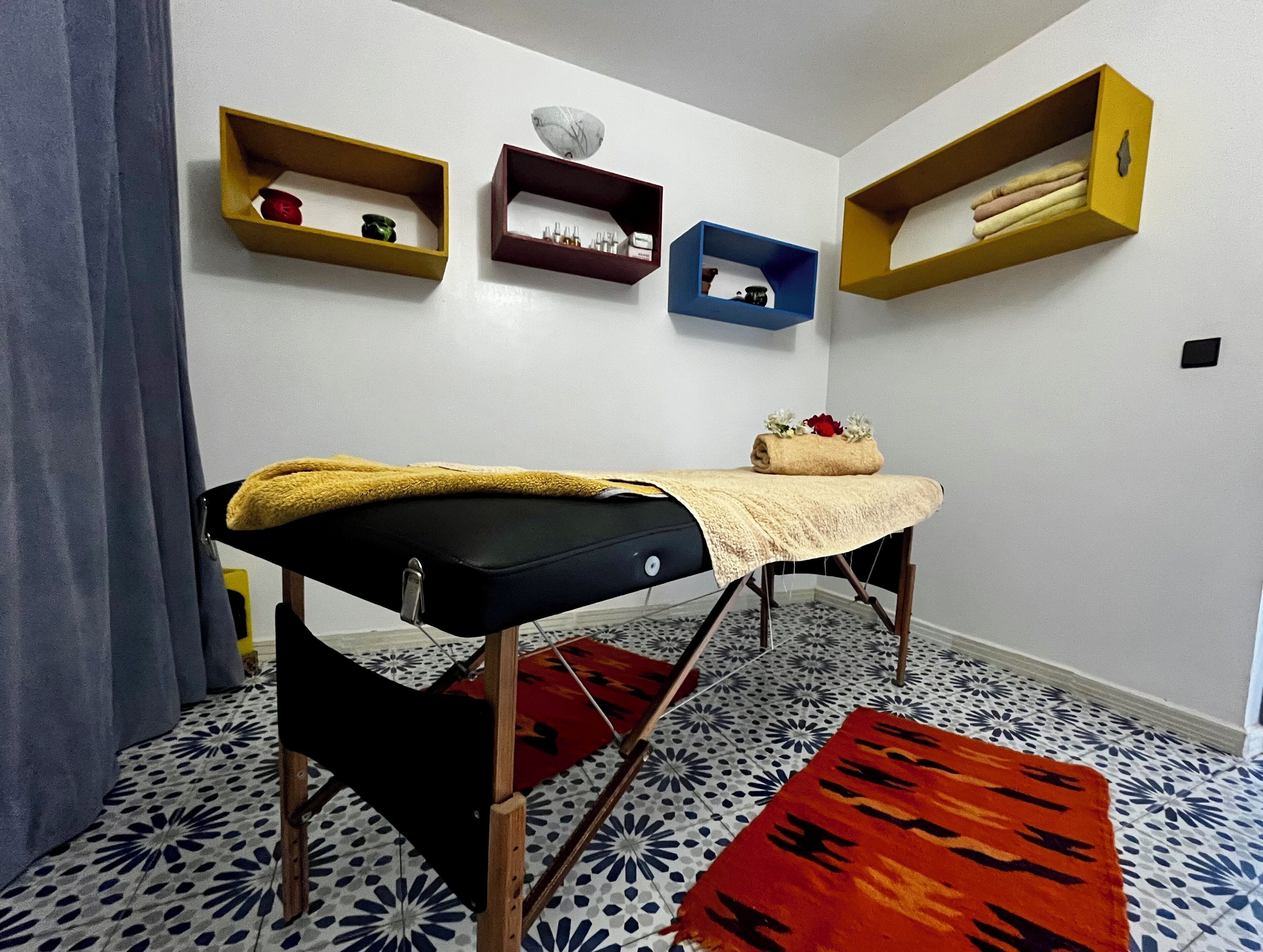 A room with a massage table, patterned floor tiles, and wall-mounted shelves with various items.