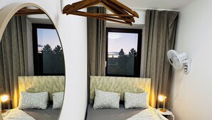 Ein Hotelzimmer mit einem großen Bett, einem runden Spiegel, einem Nachttisch mit Lampe, einem Wandventilator und Blick ins Freie durch ein Fenster.