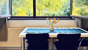 Ein Arbeitsraum mit einem großen Fenster, einem Tisch mit blauer Unterlage, zwei schwarzen Stühlen und einer Vase mit Blumen.