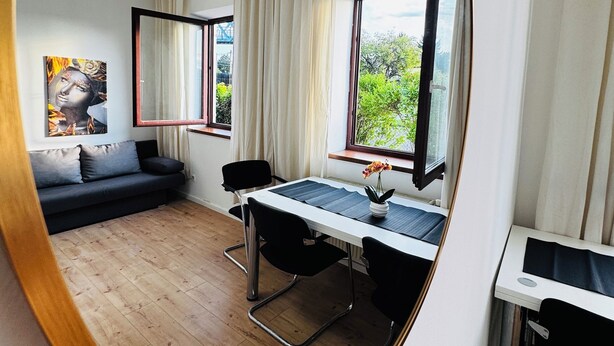 Ein modernes Zimmer mit einem Esstisch, zwei Stühlen, einem Sofa und einem großen Spiegel, der den Raum wiederspiegelt.