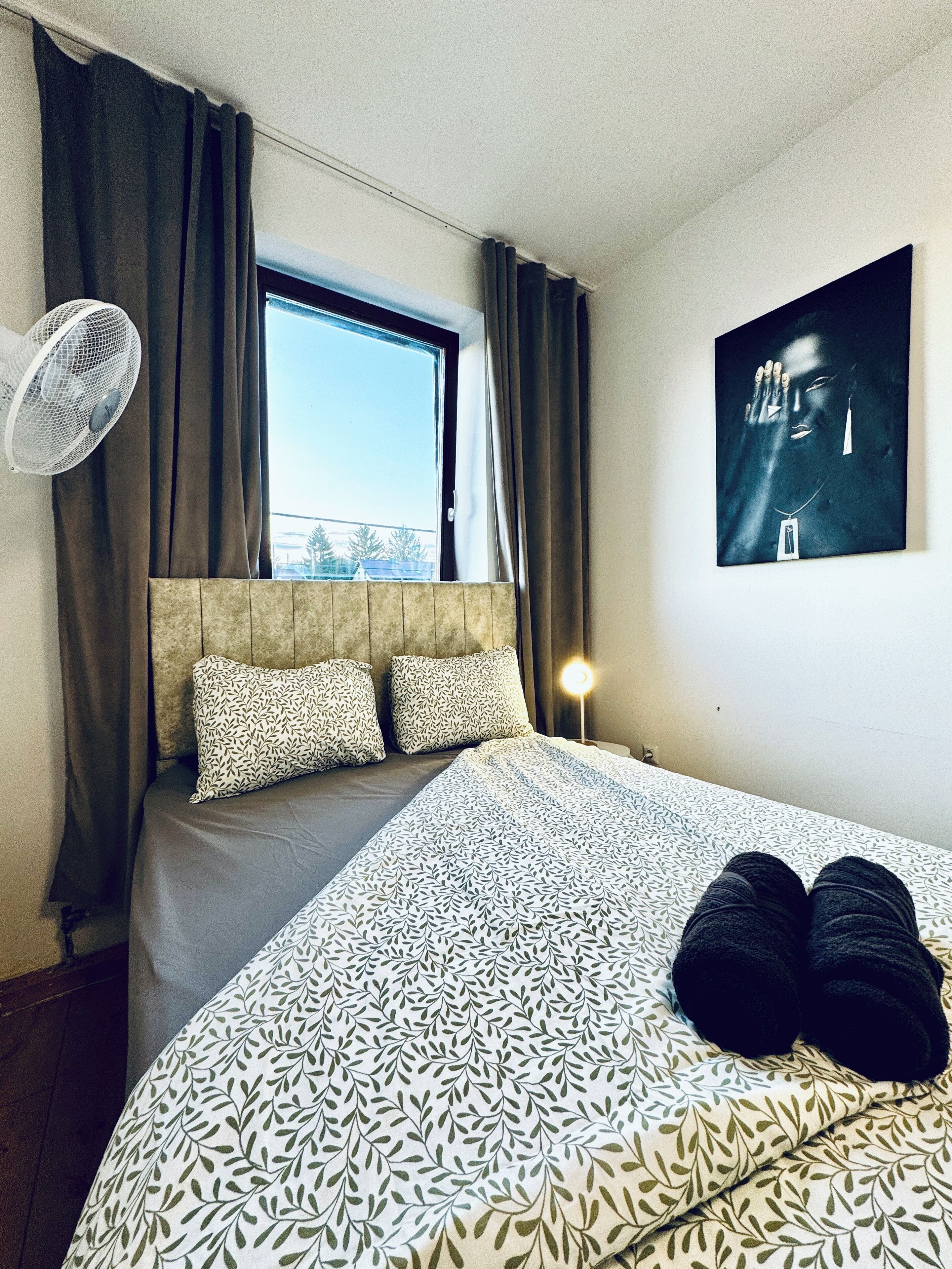 Ein Hotelzimmer mit einem Bett, einem Fenster mit Vorhängen, einem Wandventilator und einem gerahmten Bild.