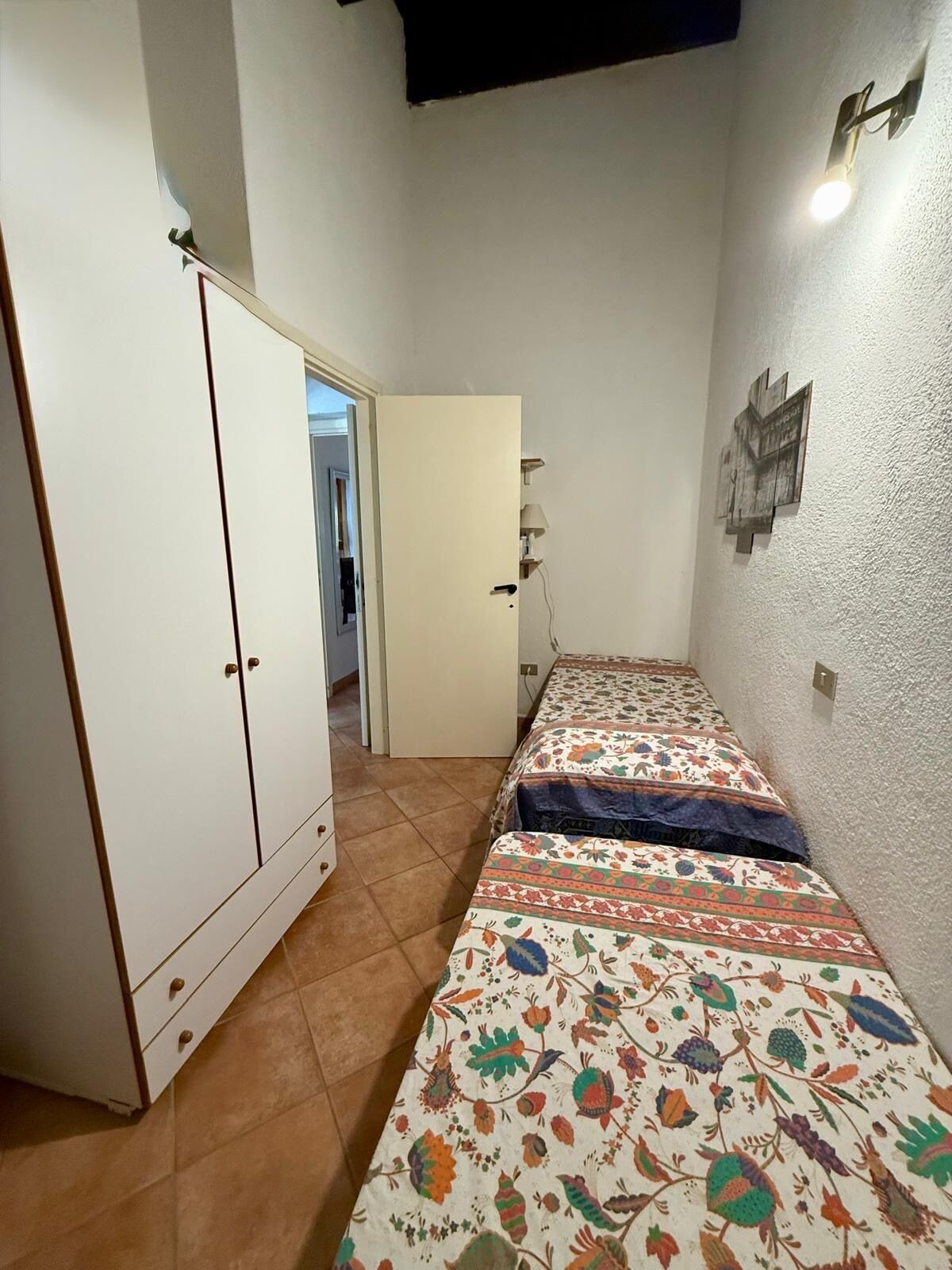 Ein schmales Zimmer mit zwei Betten, einem Kleiderschrank, einer Tür zu einem anderen Raum und einer Wandleuchte.