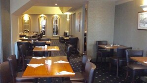 Een hotelrestaurant met meerdere tafels en stoelen, een kamerbreed tapijt, en een zicht op het restaurant op de achtergrond.