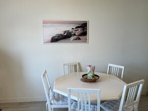 Uma área de refeições com uma mesa redonda, quatro cadeiras brancas, um vaso com flores e uma imagem emoldurada de uma praia na parede.