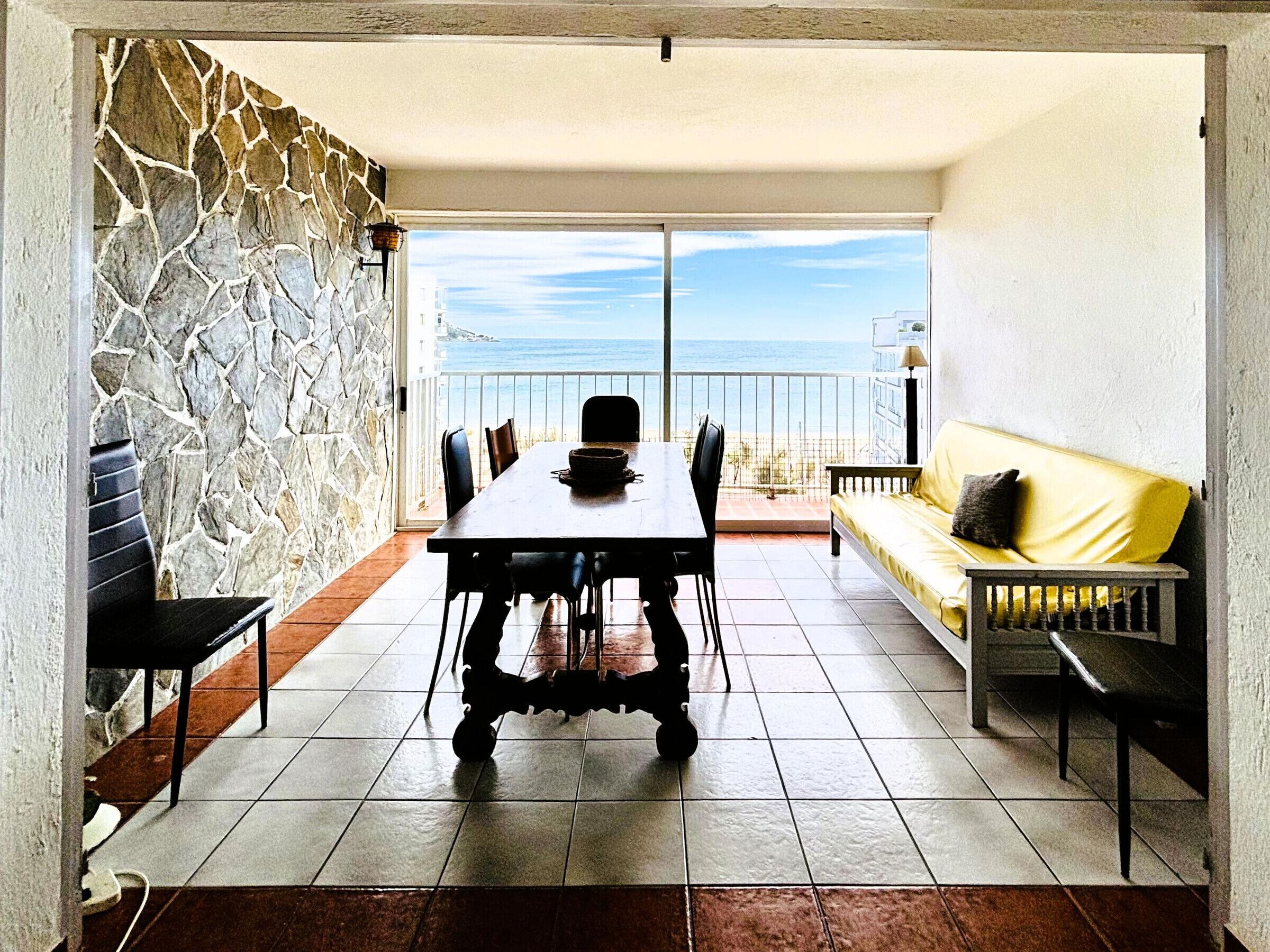 A dining area with a table and chairs, a yellow sofa, a stone wall, and a view of the sea.