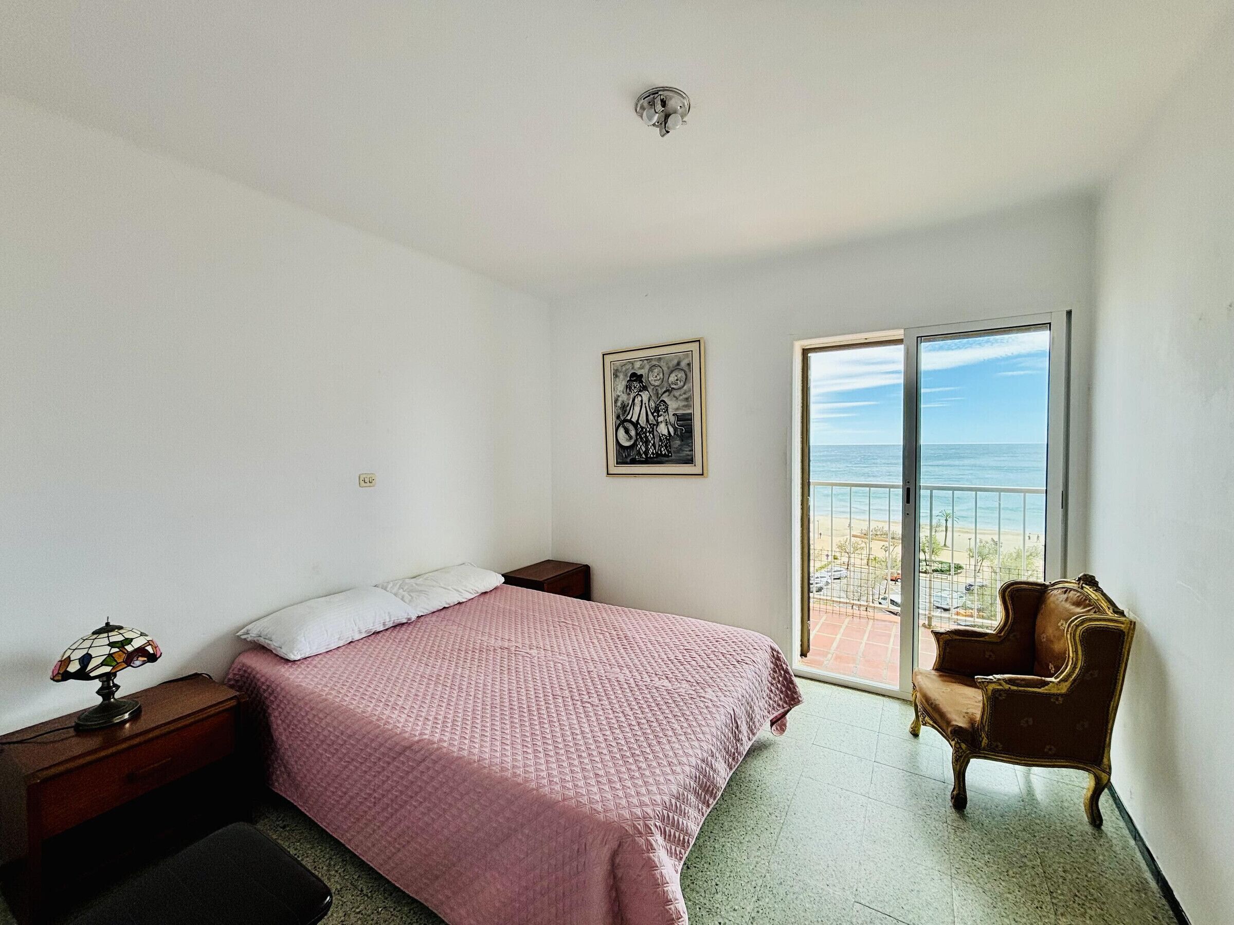 A bedroom with a bed, a chair, a picture on the wall, and a balcony with a view.