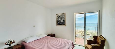 A bedroom with a bed, a chair, a picture on the wall, and a balcony with a view.