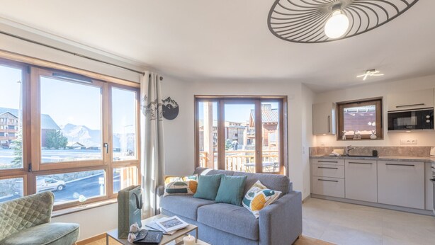 Una sala moderna con un sofá, una mesita y una silla. Hay una zona de cocina con microondas y armarios. Amplias ventanas ofrecen vistas a edificios y montañas.