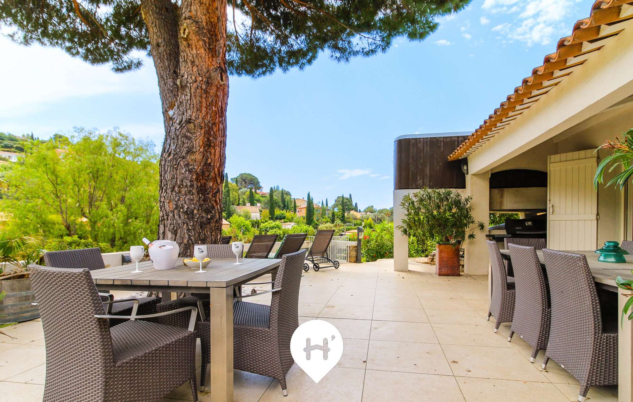 An outdoor patio area with wicker furniture, a dining table, and a view of a lush garden and hills in the background.