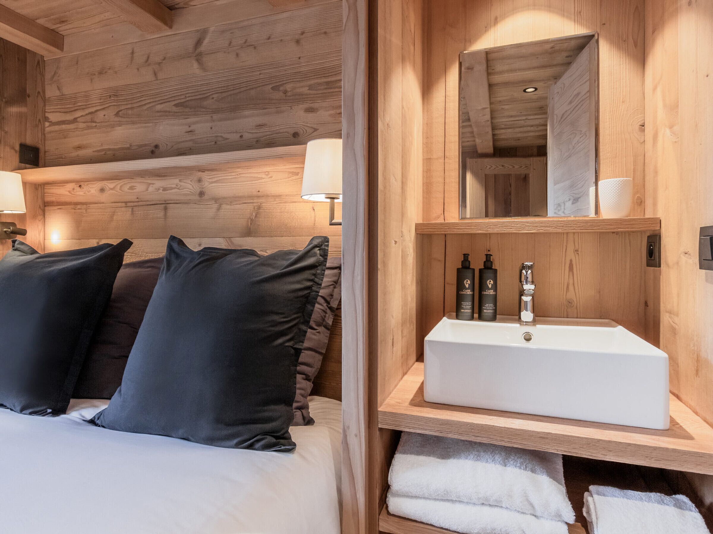 A wooden cabin room with a bed, pillows, a bedside table with a lamp, a mirror, and a shelf with toiletries.