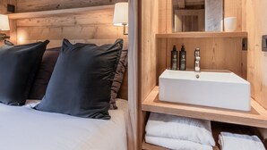 A wooden cabin room with a bed, pillows, a bedside table with a lamp, a mirror, and a shelf with toiletries.