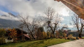 Une terrasse en bois menant à une maison dotée d’un toit en pente, entourée d’arbres et avec une chaîne de montagnes en arrière-plan.