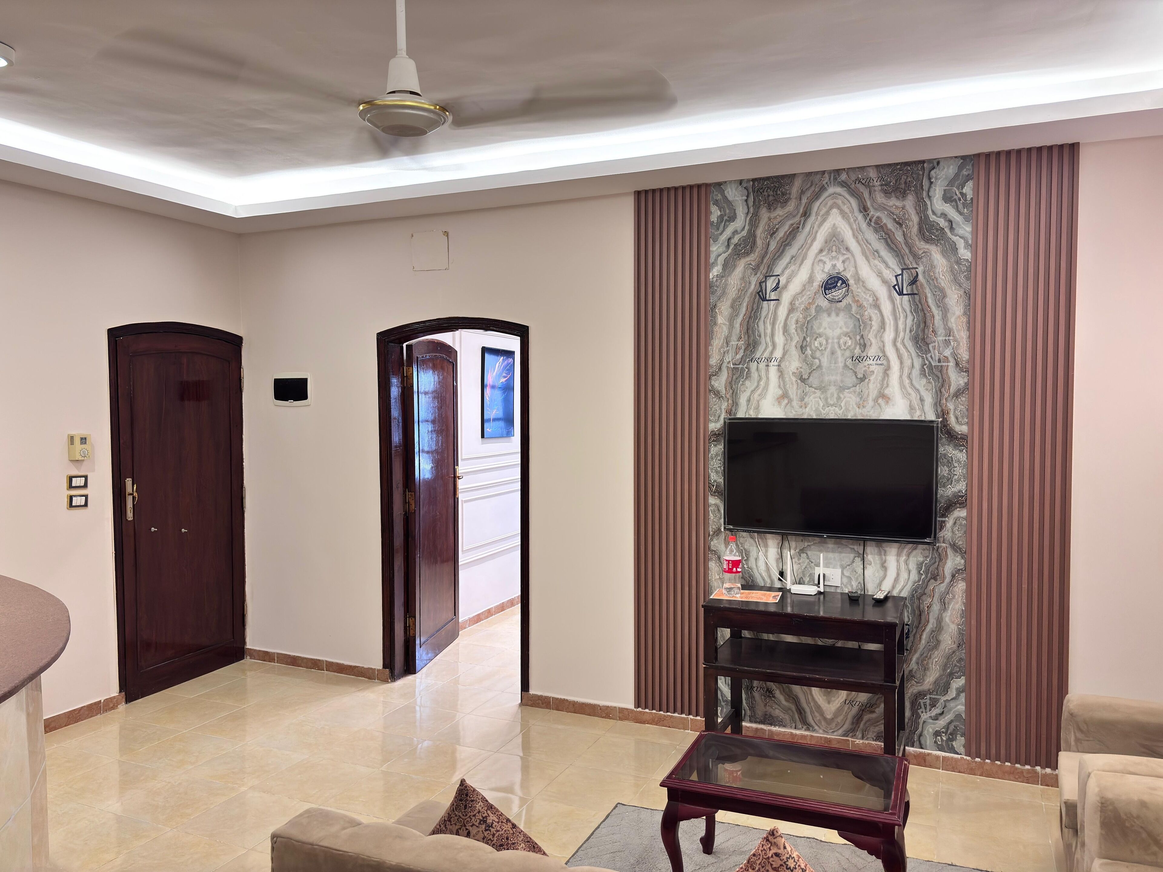 A modern living room with a flat-screen TV mounted on a marble wall, a wooden door, and a ceiling fan.