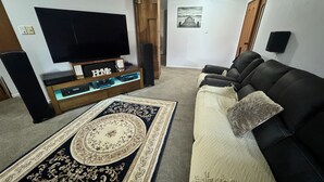 Un salon avec un canapé en cuir noir, un grand téléviseur à écran plat posé sur un meuble en bois, un tapis à motifs et un haut-parleur fixé au mur.