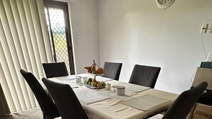 Un coin repas avec une table dressée pour quatre personnes, entourée de chaises, une horloge au mur et une fenêtre avec des stores.