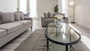 Een moderne woonkamer met een glazen salontafel, grijze banken en een potplant.