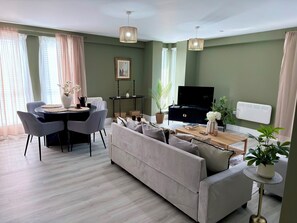 Ein Wohnzimmer mit einer Couch, einem Esstisch mit Stühlen, einem Fernseher auf einem Schrank und einer Heizung.