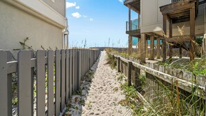 A sandy pathway flanked by wooden fences leading to a building with balconies and an air conditioning unit. - "Forever Summer" | Dog Friendly | Pensacola Beach | My Beach Getaways (Pensacola Beach)