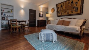 Uma sala de estar com sofá, mesa de jantar com cadeiras, estante de livros e um grande tapeçaria decorativa na parede.