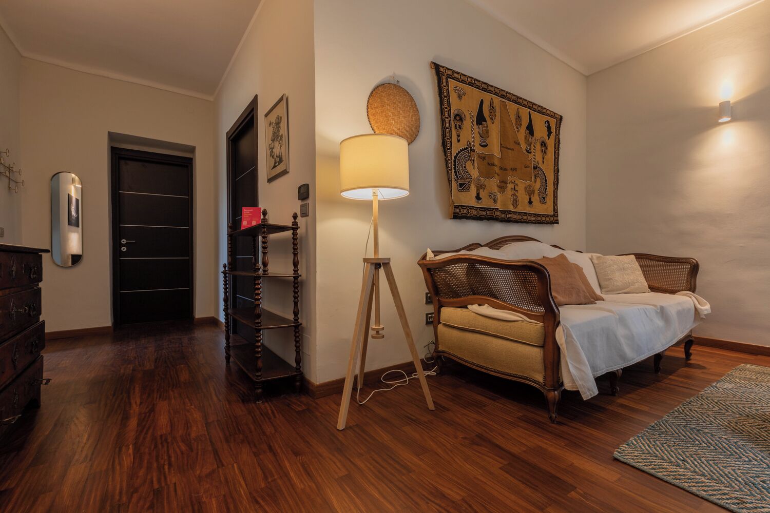 A bedroom with a wooden floor, a bed with a headboard, a standing lamp, a wall-mounted picture, and a door.