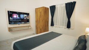 Una habitación de hotel con cama, televisor montado en la pared, armario de madera y ventana con cortinas.