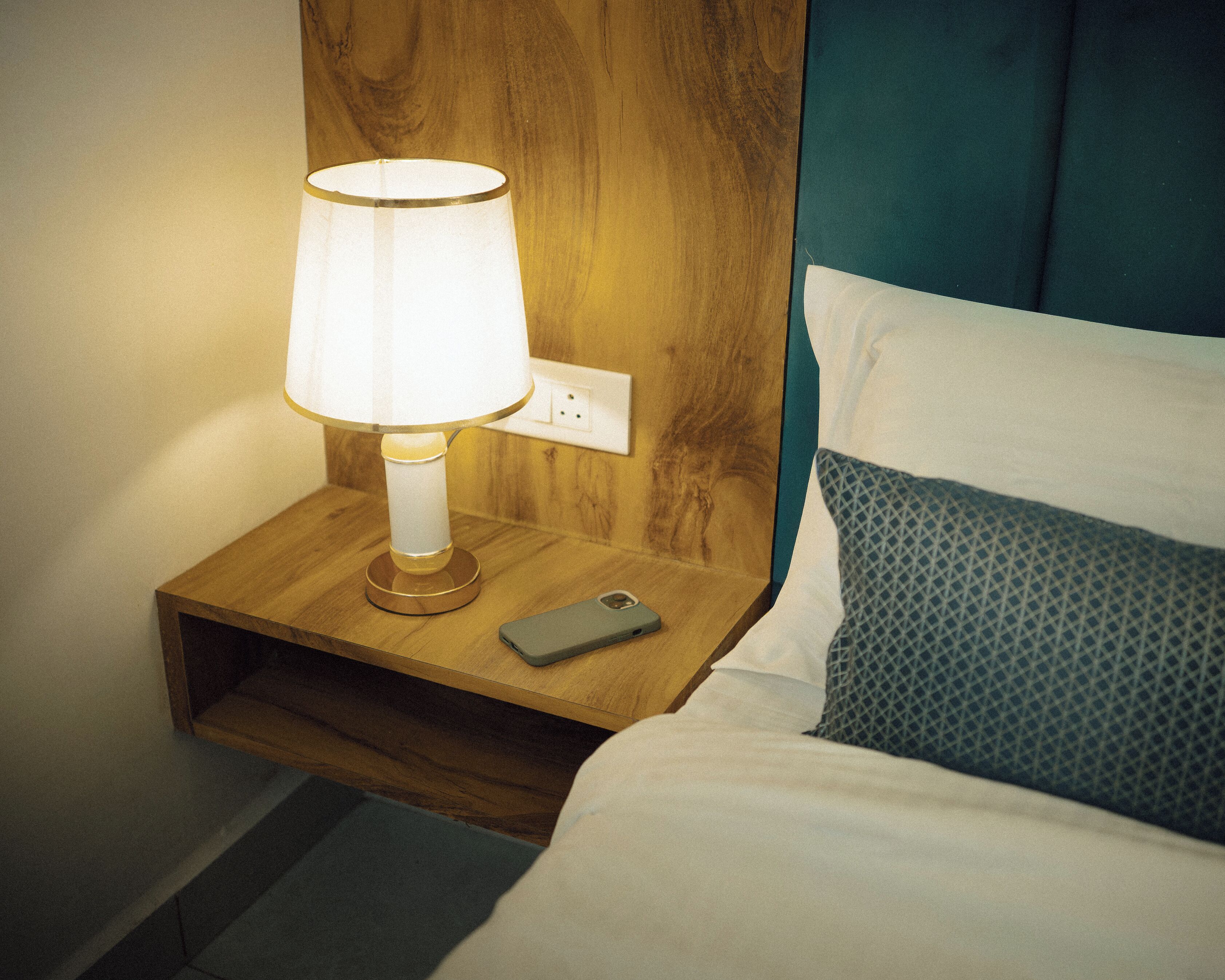 A wooden bedside table with a lamp, a smartphone, and a patterned pillow on a bed.